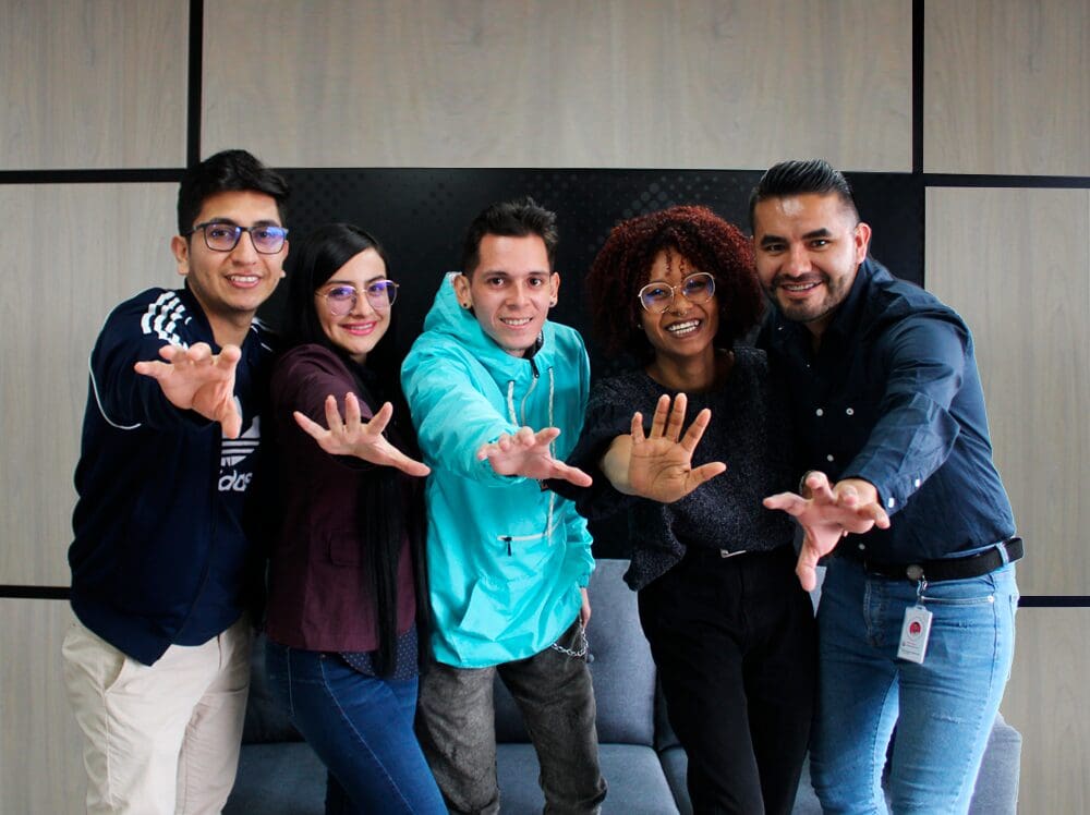 Cinco colegas sonrientes posando con las manos extendidas hacia la cámara en una oficina.